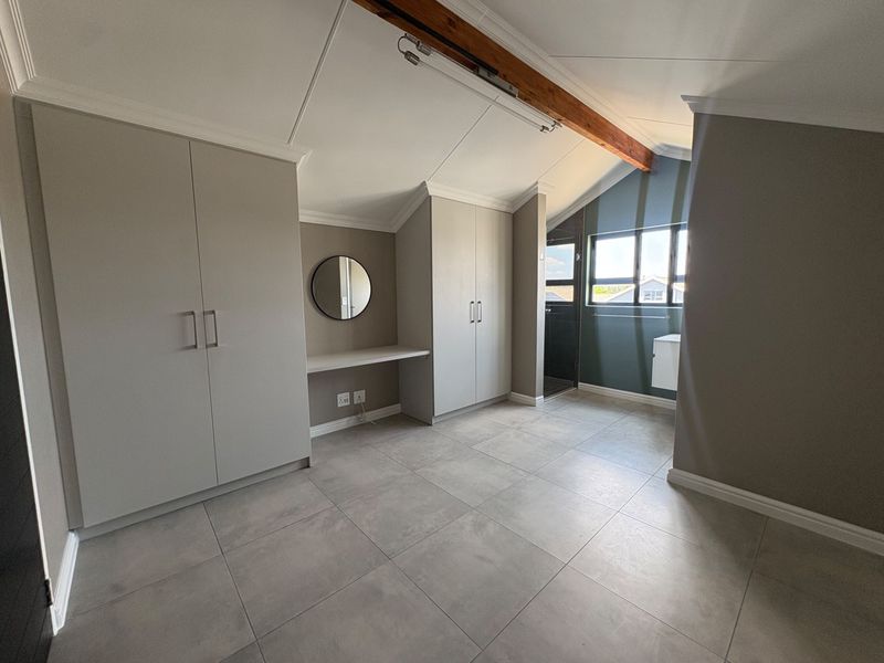 A bright, modern bedroom featuring vaulted ceilings with an exposed timber beam, neutral tiled flooring, and ample built-in cupboard space with a vanity area.