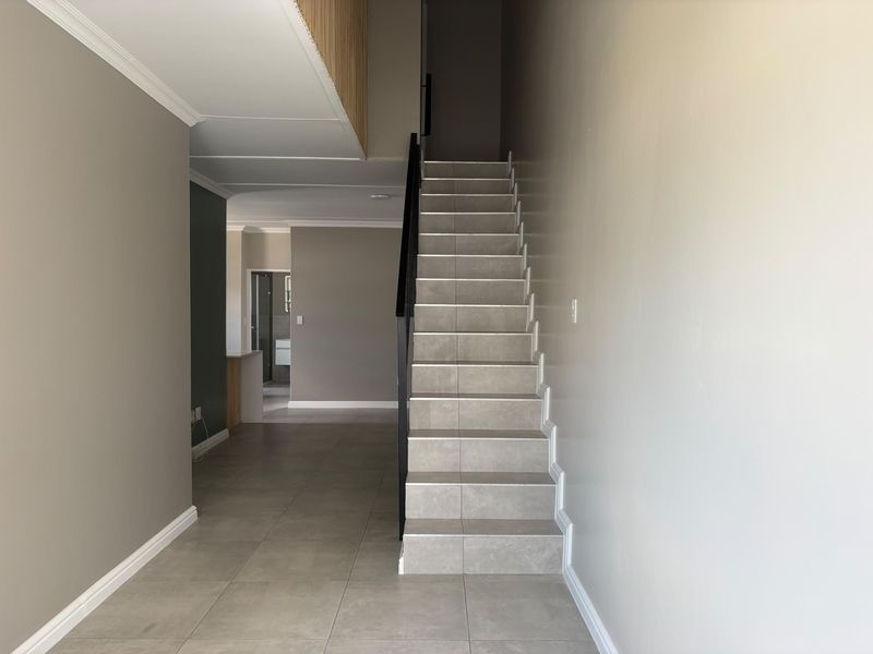A bright, modern hallway featuring neutral wall tones, ceramic floor tiles, and a clean staircase with a minimalist metal handrail.