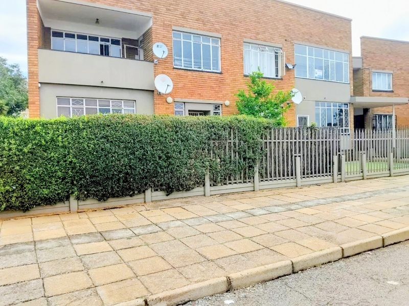 Front exterior view of a multi-story brick apartment building featuring a paved sidewalk, security fencing, and established hedging.