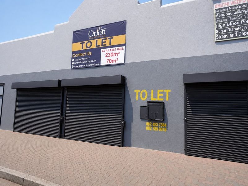 Commercial storefront featuring multiple roller shutter doors, paved frontage, and prominent signage indicating available retail or office space for lease.