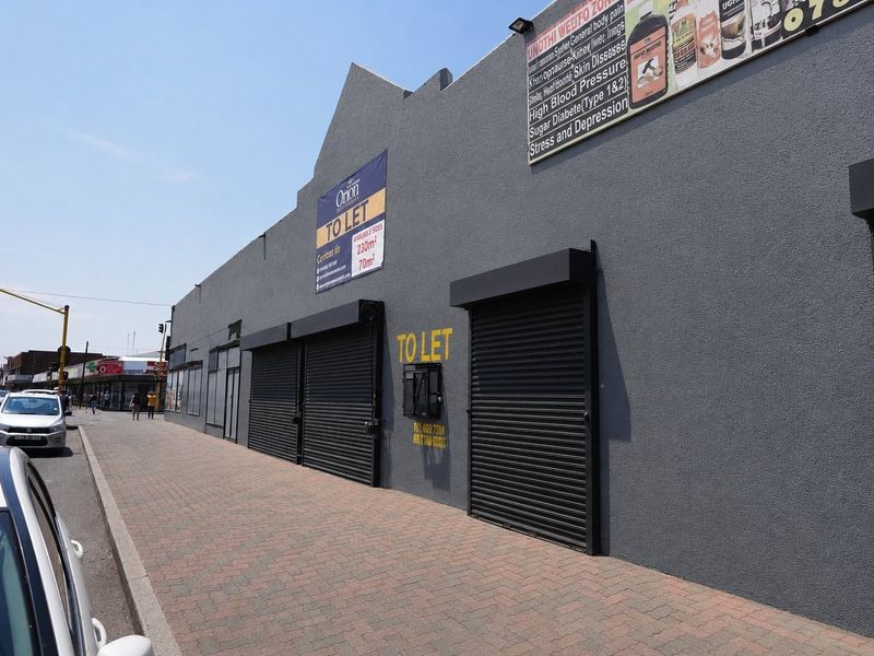 Commercial street-front facade featuring multiple roller shutter doors, paved sidewalk access, and prominent signage for available retail or office space.