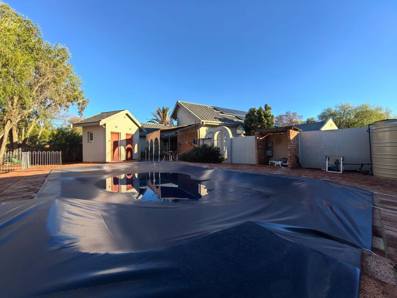 Outdoor pool area featuring a covered swimming pool, brick paving, and a water storage tank, set against a residential outbuilding and garden.