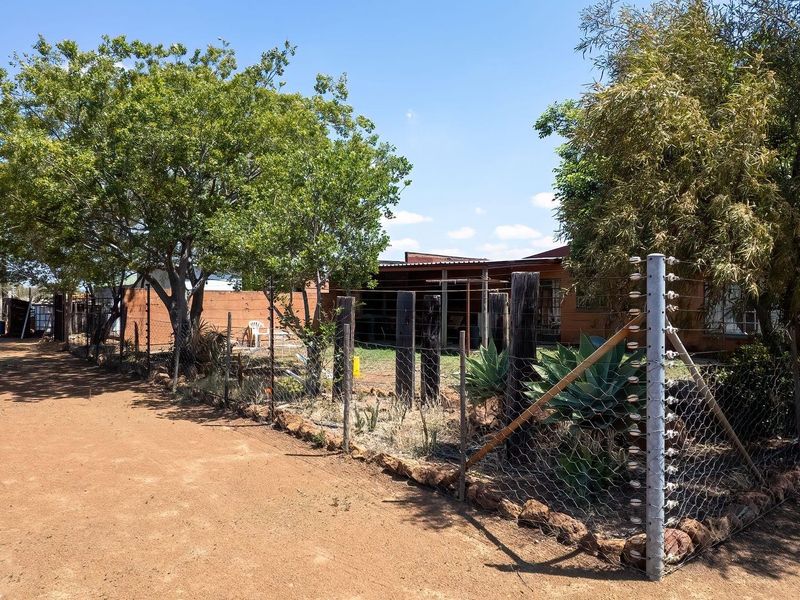 Front view of a farm property featuring a dirt driveway, established trees, and perimeter security fencing with an electric wire system.