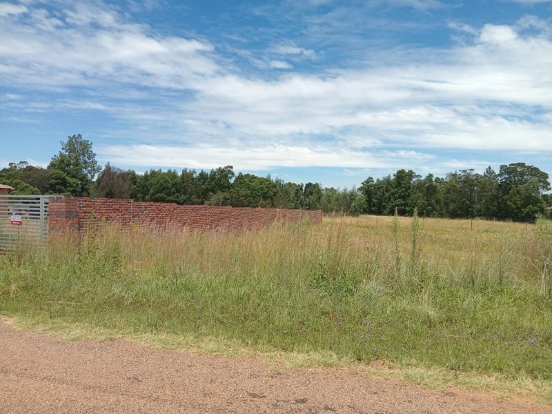 A vacant residential plot featuring a brick boundary wall and metal gate, surrounded by natural grass and mature trees.