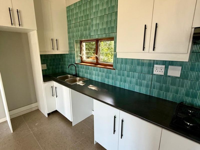 A bright, modern kitchen featuring white cabinetry, a striking green tiled backsplash, black countertops, and a built-in hob, offering a clean and functional space.