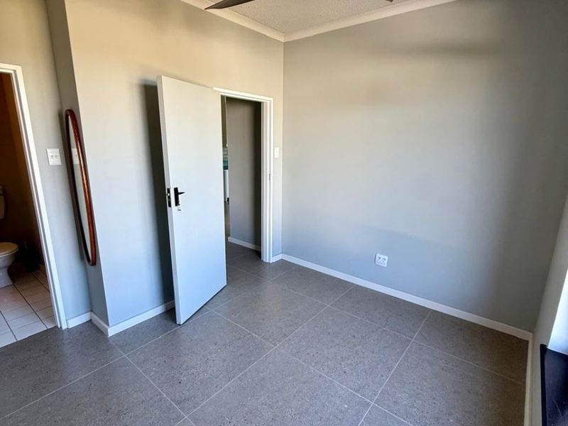 A clean, empty bedroom featuring modern grey floor tiles, a ceiling fan, and neutral wall paint, with an adjacent bathroom visible through the doorway.