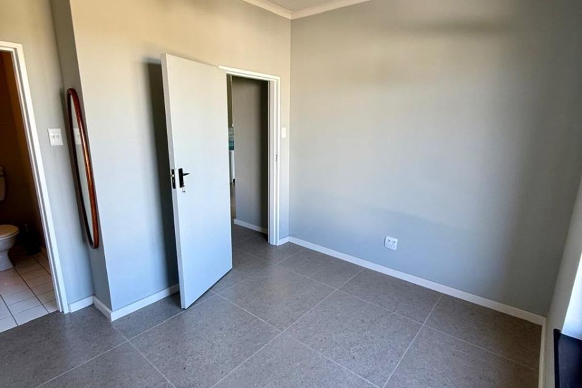 A clean, empty bedroom featuring modern grey floor tiles, a ceiling fan, and neutral wall paint, with an adjacent bathroom visible through the doorway.