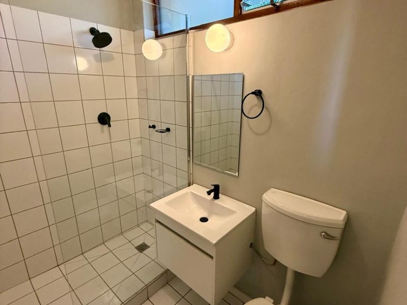 A modern, compact bathroom featuring a walk-in shower with glass screen, wall-mounted vanity, and louver windows for natural ventilation.