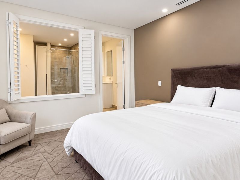 A well-appointed bedroom featuring neutral tones, recessed lighting, carpeted flooring, and an internal window providing a view into the en-suite bathroom.