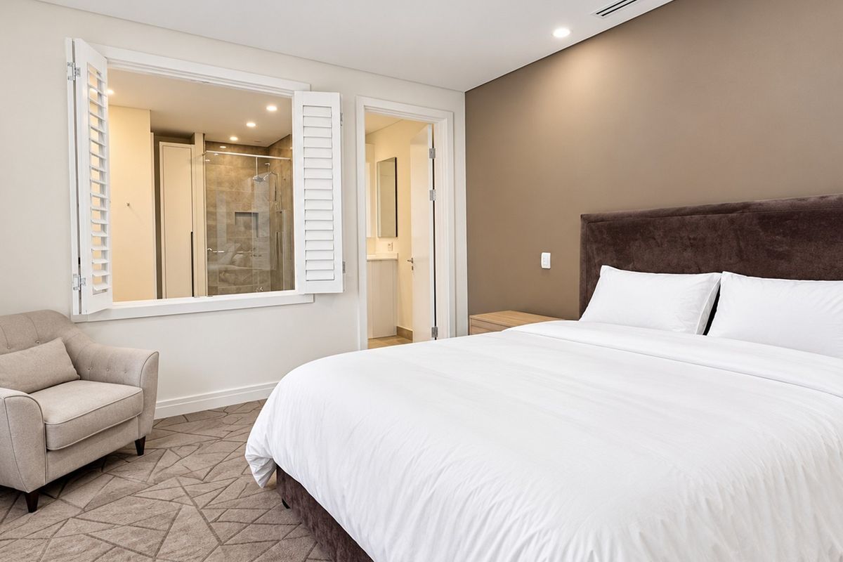 A well-appointed bedroom featuring neutral tones, recessed lighting, carpeted flooring, and an internal window providing a view into the en-suite bathroom.