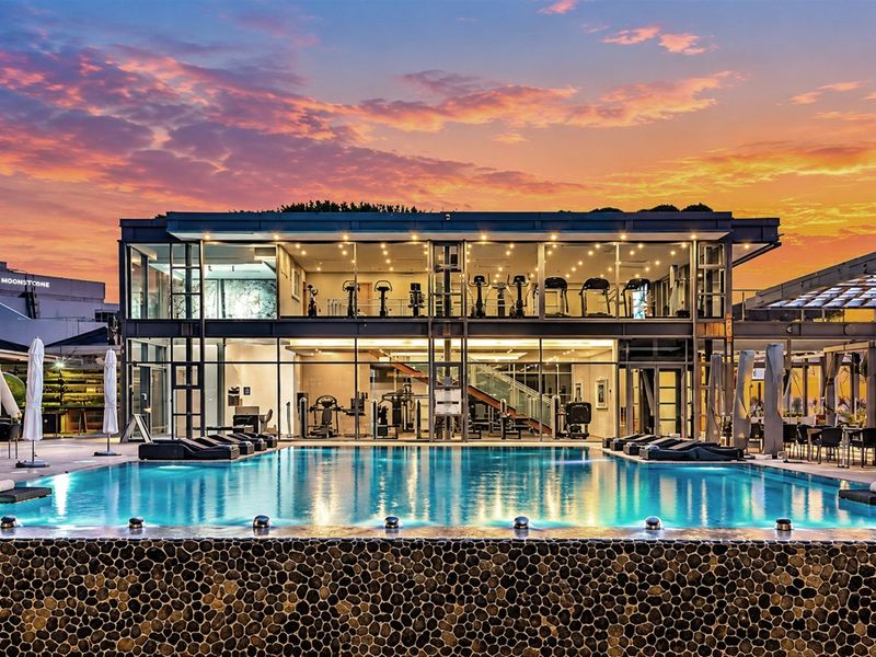 A modern apartment complex pool area featuring a stone-clad infinity edge pool, expansive glass facade, and integrated gym facilities at sunset.