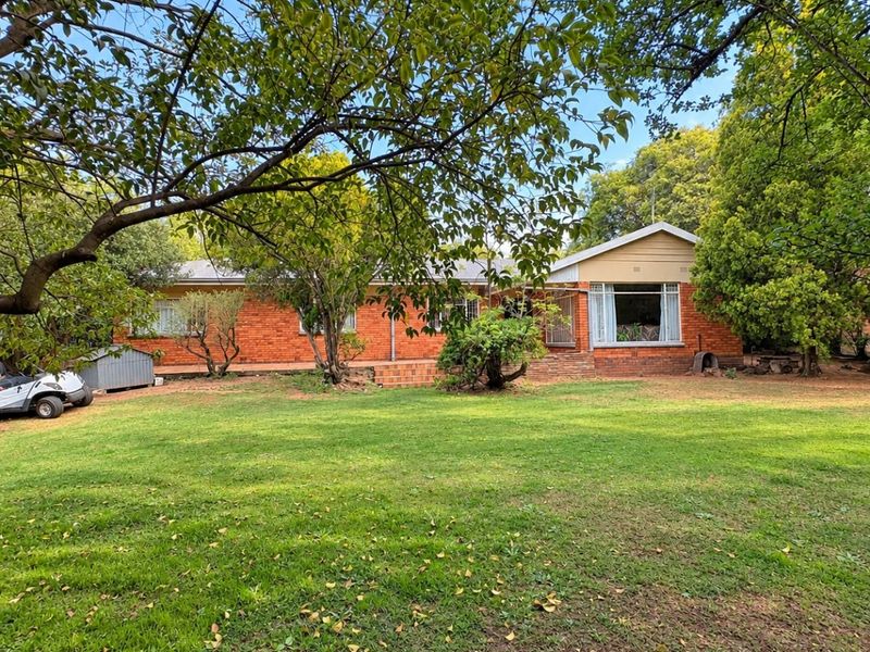 A spacious rear garden view of a single-story brick house featuring a large lawn, mature trees, and a paved patio area.