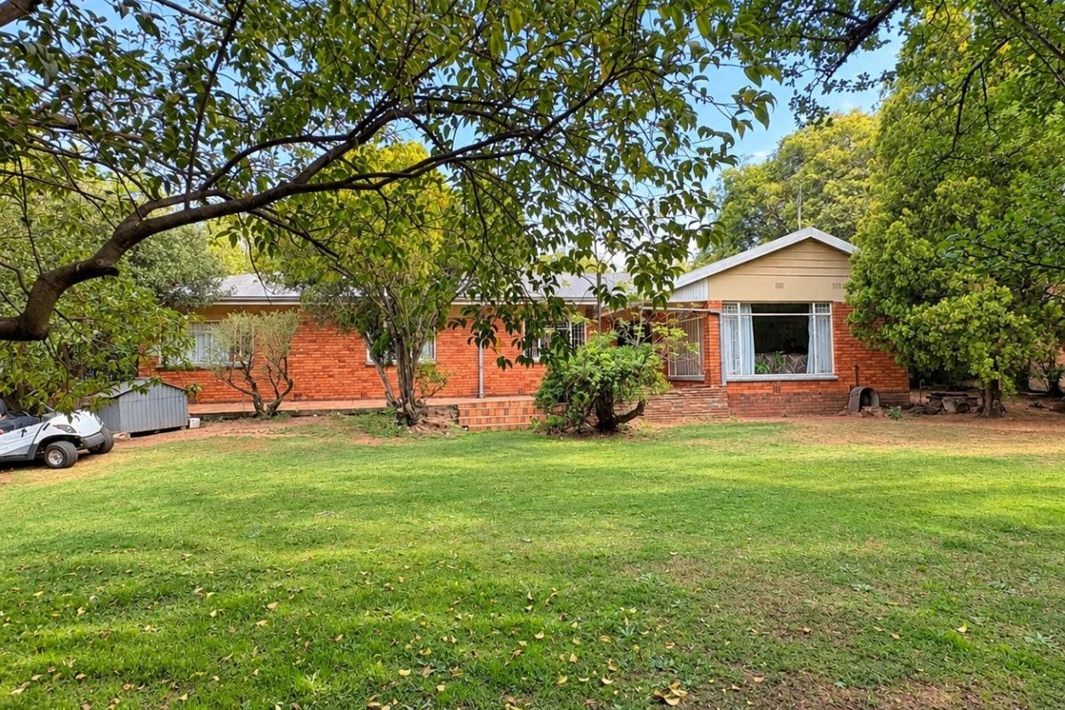 A spacious rear garden view of a single-story brick house featuring a large lawn, mature trees, and a paved patio area.