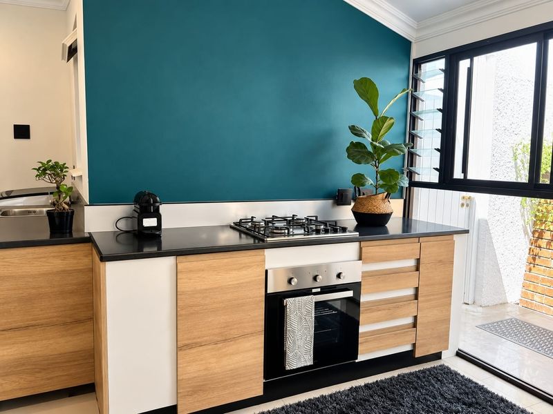 Modern kitchen featuring wood-tone cabinetry, black stone countertops, a gas hob, and a built-in oven, set against a bold teal accent wall.