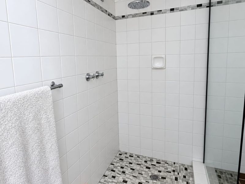 A clean, modern walk-in shower featuring white ceramic wall tiles, a mosaic tiled floor, a rain shower head, and a glass screen.