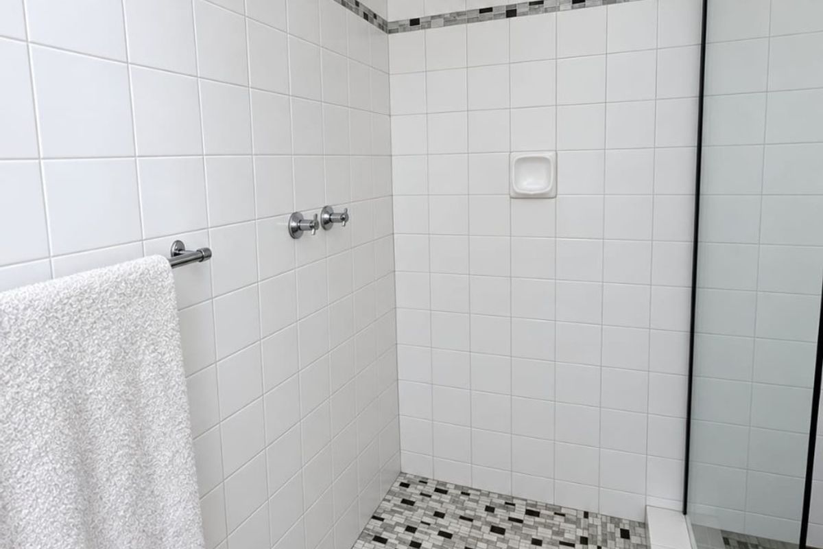 A clean, modern walk-in shower featuring white ceramic wall tiles, a mosaic tiled floor, a rain shower head, and a glass screen.