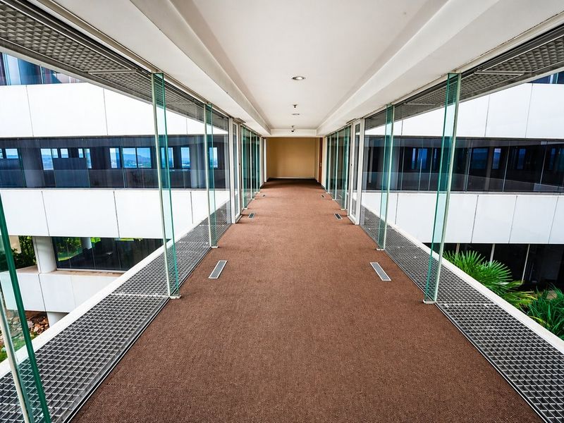 A modern, elevated glass-enclosed walkway connecting office building sections, featuring clean lines, carpeted flooring, and integrated metal ventilation grilles.
