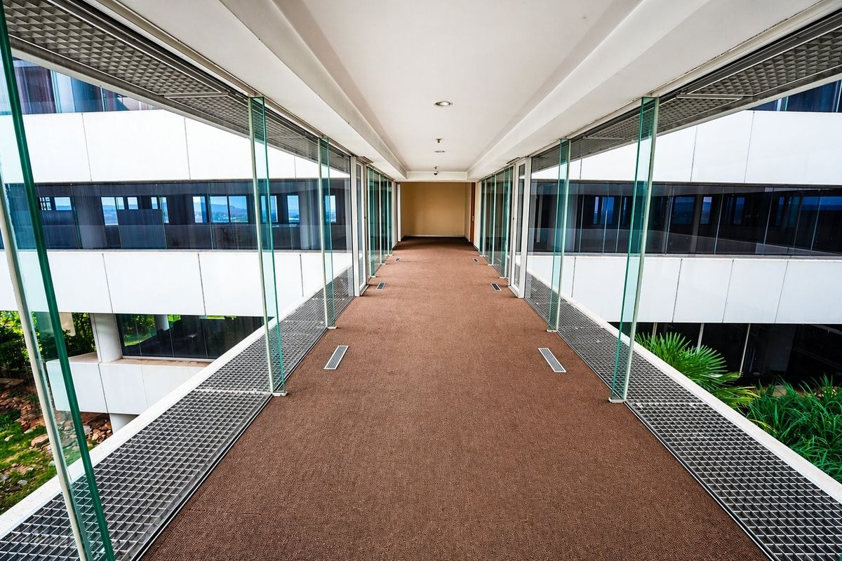A modern, elevated glass-enclosed walkway connecting office building sections, featuring clean lines, carpeted flooring, and integrated metal ventilation grilles.