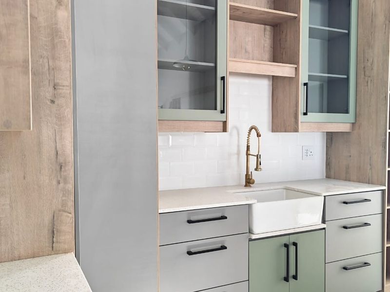 Modern kitchen featuring a farmhouse sink, brass faucet, subway tile backsplash, and a mix of wood-tone and sage green cabinetry with stone countertops.
