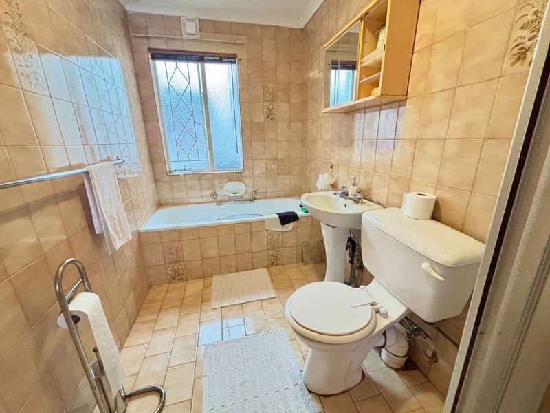 A dated bathroom featuring a bathtub, pedestal basin, and toilet with neutral wall and floor tiling.