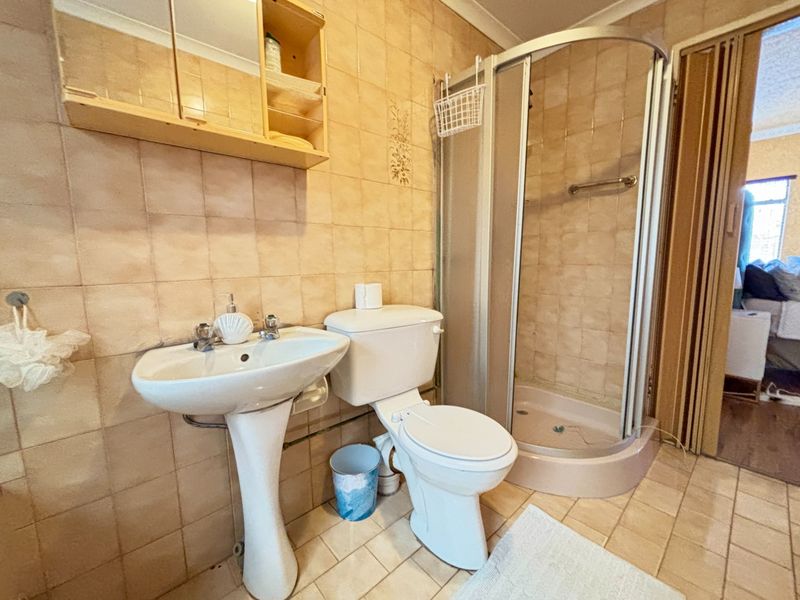 A functional bathroom featuring a pedestal basin, toilet, and corner shower enclosure with dated wall and floor tiling throughout.