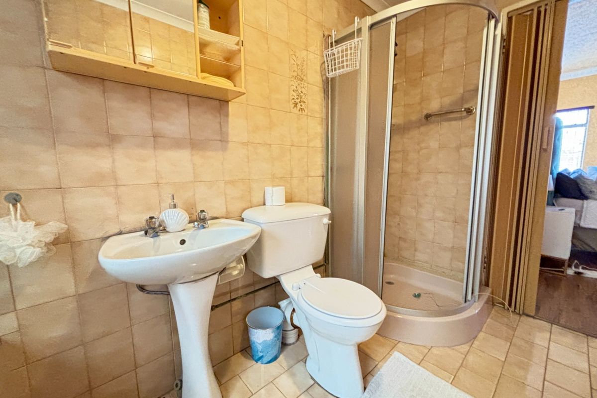 A functional bathroom featuring a pedestal basin, toilet, and corner shower enclosure with dated wall and floor tiling throughout.