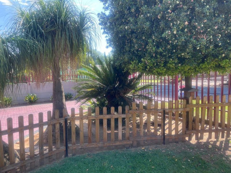 A view of the front garden area featuring a lawn, wooden picket fencing, paved driveway, and established trees with perimeter security fencing.