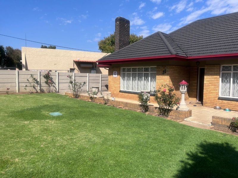 A well-maintained rear exterior view of a facebrick house featuring a tiled roof, brick chimney, and a spacious lawn with a paved patio area.