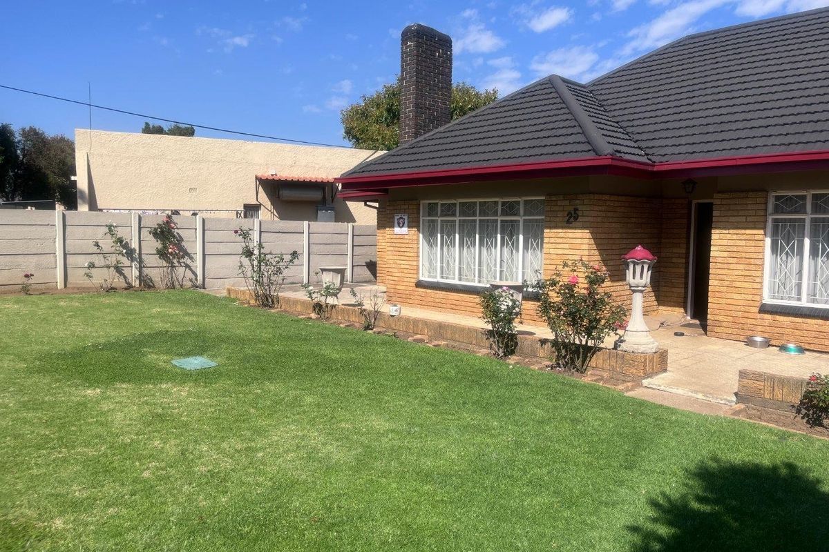 A well-maintained rear exterior view of a facebrick house featuring a tiled roof, brick chimney, and a spacious lawn with a paved patio area.