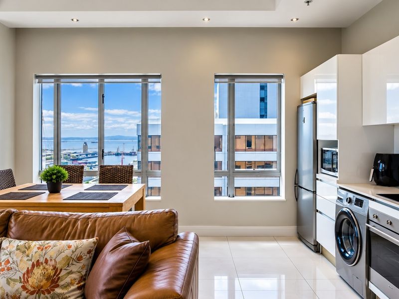 Modern open-plan living and kitchen area featuring high-gloss cabinetry, integrated appliances, tiled floors, and large windows with expansive harbour views.