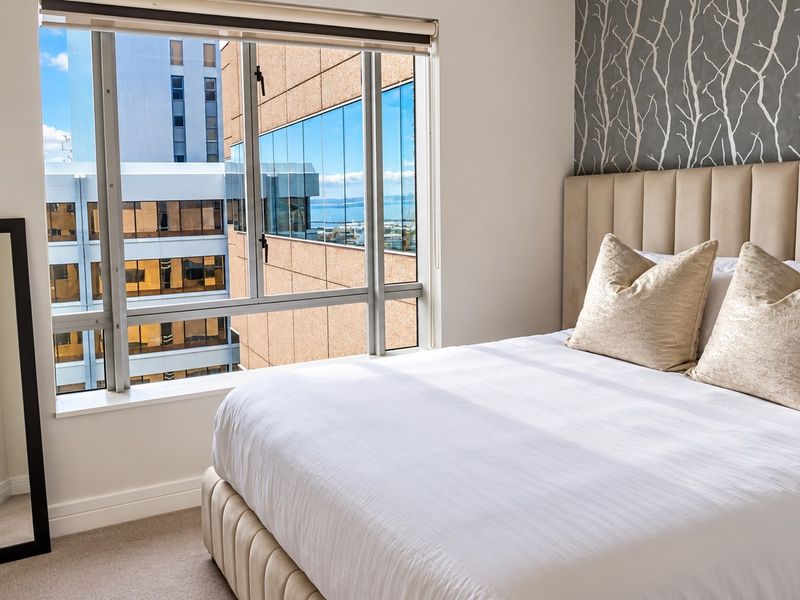 A well-maintained bedroom featuring a large window with city views, neutral carpet, and a stylish upholstered headboard with feature wallpaper.