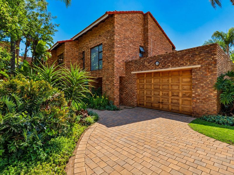 A well-maintained face brick house featuring a paved driveway, a wooden garage door, and established garden landscaping.