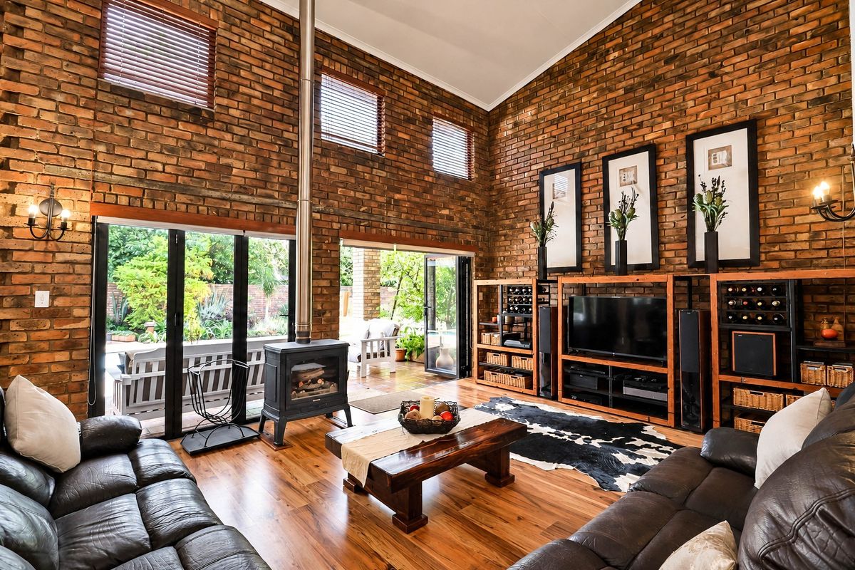 Spacious living room featuring high vaulted ceilings, exposed brick walls, timber flooring, and a central wood-burning fireplace with direct access to the patio.