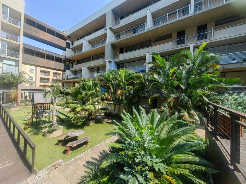 A well-maintained communal garden area featuring lush landscaping, paved pathways, and wooden decking, set against the modern concrete facade of the apartment complex.