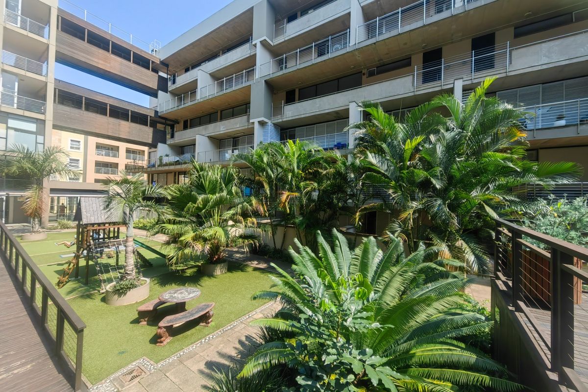 A well-maintained communal garden area featuring lush landscaping, paved pathways, and wooden decking, set against the modern concrete facade of the apartment complex.