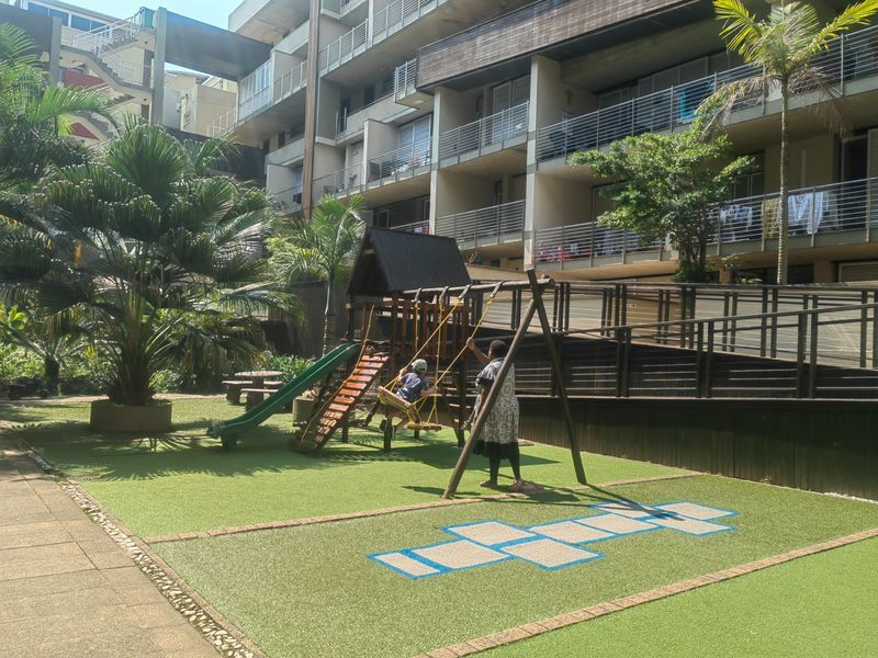 Communal outdoor area featuring a playground with artificial turf, paved pathways, and access ramps leading to the apartment building balconies.