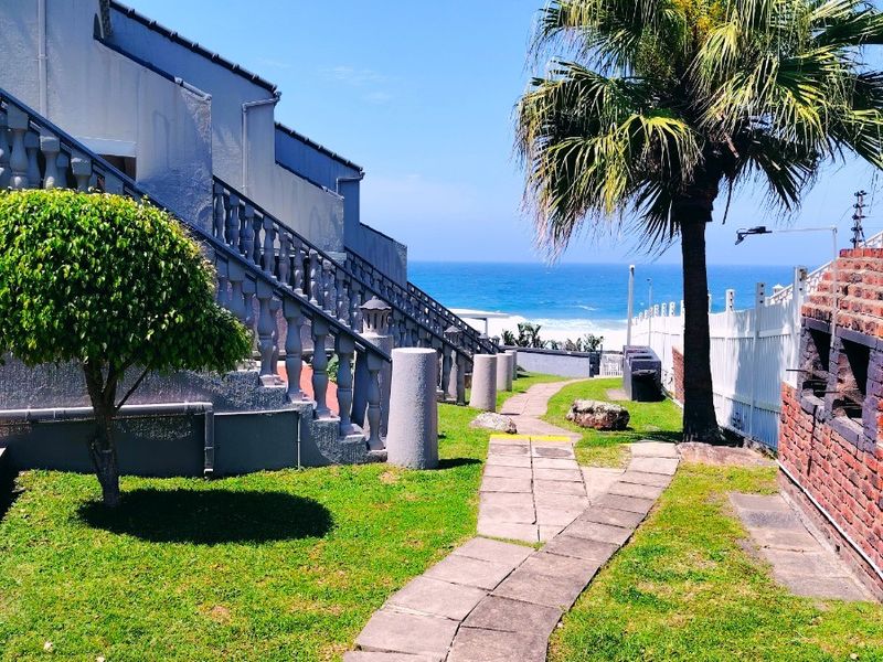 Rear garden area featuring a paved walkway, built-in brick braai, and concrete balustrade staircase, with ocean views in the background.