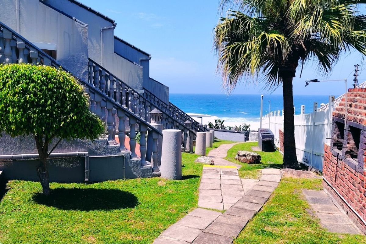 Rear garden area featuring a paved walkway, built-in brick braai, and concrete balustrade staircase, with ocean views in the background.