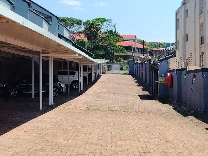 A paved driveway leading to a row of covered parking bays within an apartment complex, featuring perimeter security walls and electric fencing.