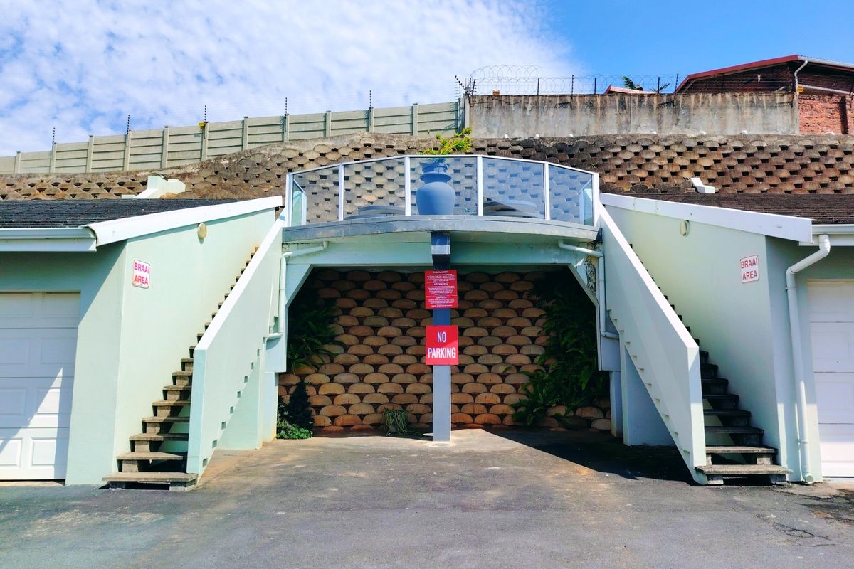 Exterior view of the property featuring a raised balcony with glass balustrade, concrete staircases, and integrated garage access with a retaining wall backdrop.