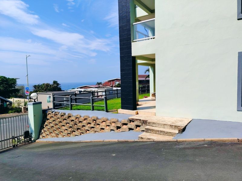 Exterior view of the apartment building entrance featuring a retaining wall, concrete steps, and a balcony with a glass balustrade overlooking the neighborhood.