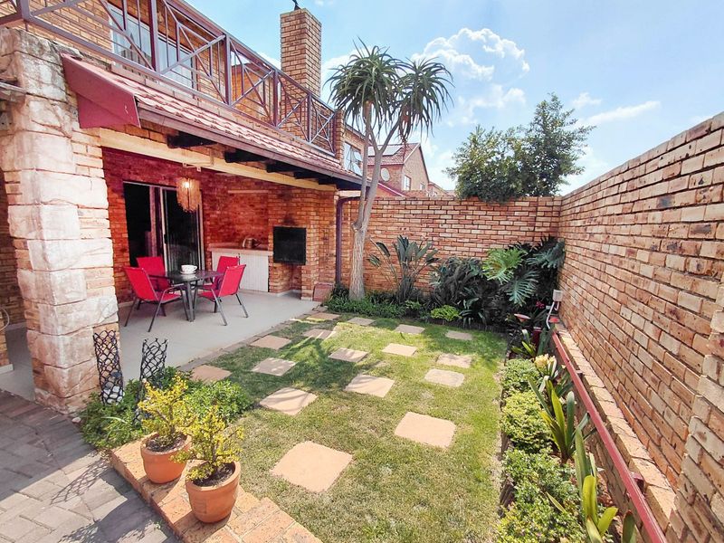 A private rear garden featuring a covered brick patio with a built-in braai, overlooking a landscaped lawn area enclosed by high brick walls.