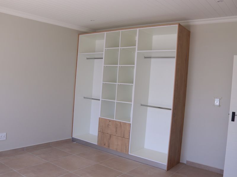 A bright bedroom featuring a modern, spacious built-in wardrobe with integrated shelving and hanging rails, complemented by neutral tiled flooring and a clean finish.