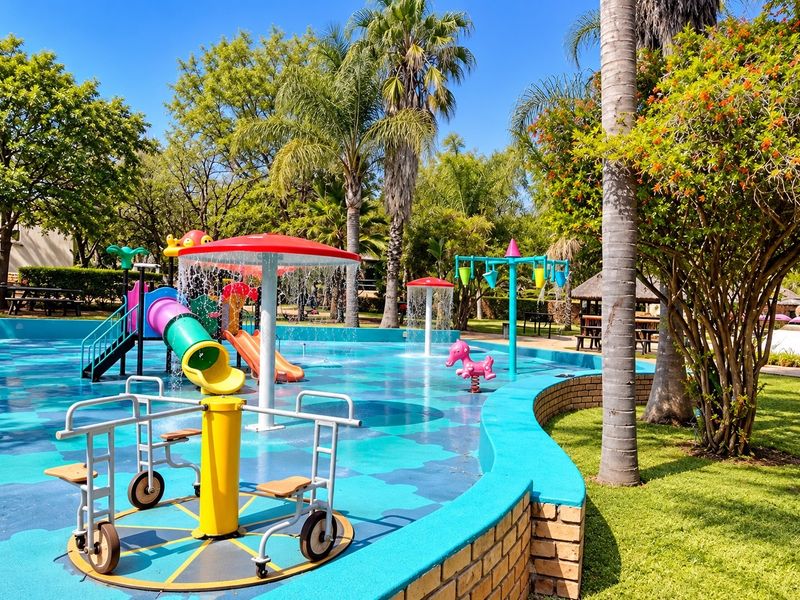 A vibrant outdoor splash pad and water play area featuring colorful slides, water features, and a surrounding landscaped garden with brick retaining walls.