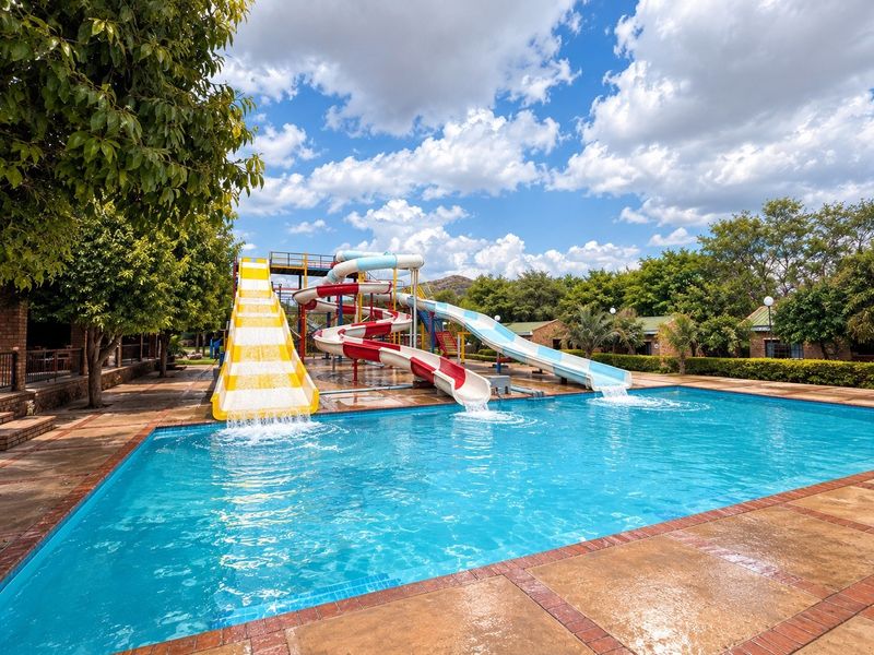 Outdoor swimming pool area featuring multiple water slides and a paved deck, set within a landscaped lodge environment.