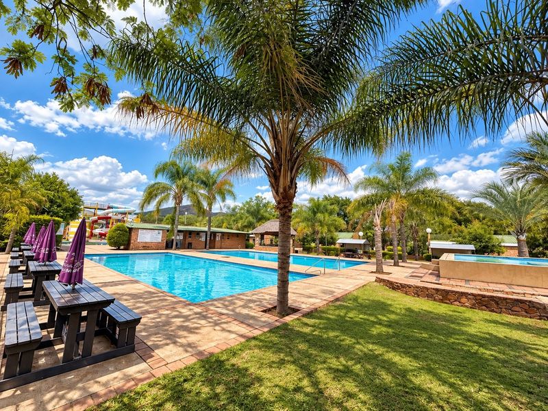 A well-maintained outdoor pool area featuring multiple swimming pools, stone retaining walls, paved surrounds, and a water slide set within a landscaped garden.