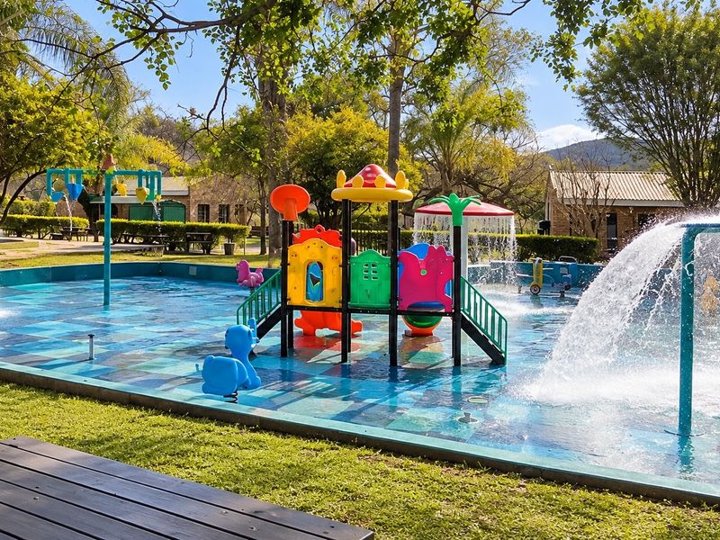 A vibrant outdoor splash pad area featuring integrated water fountains and colorful play structures, set within a well-maintained garden environment.