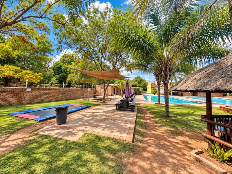 Outdoor recreational area featuring a swimming pool, paved patio, shade sail, and a traditional thatch lapa set within a landscaped garden.