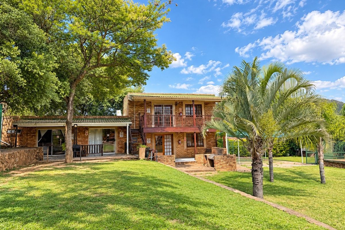 Rear view of a two-story face brick lodge featuring a timber balcony, paved patio area with a built-in brick braai, and landscaped garden.
