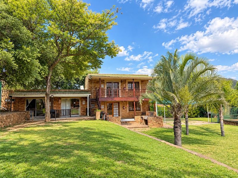 Rear view of a two-story face brick lodge featuring a timber balcony, paved patio area with a built-in brick braai, and landscaped garden.